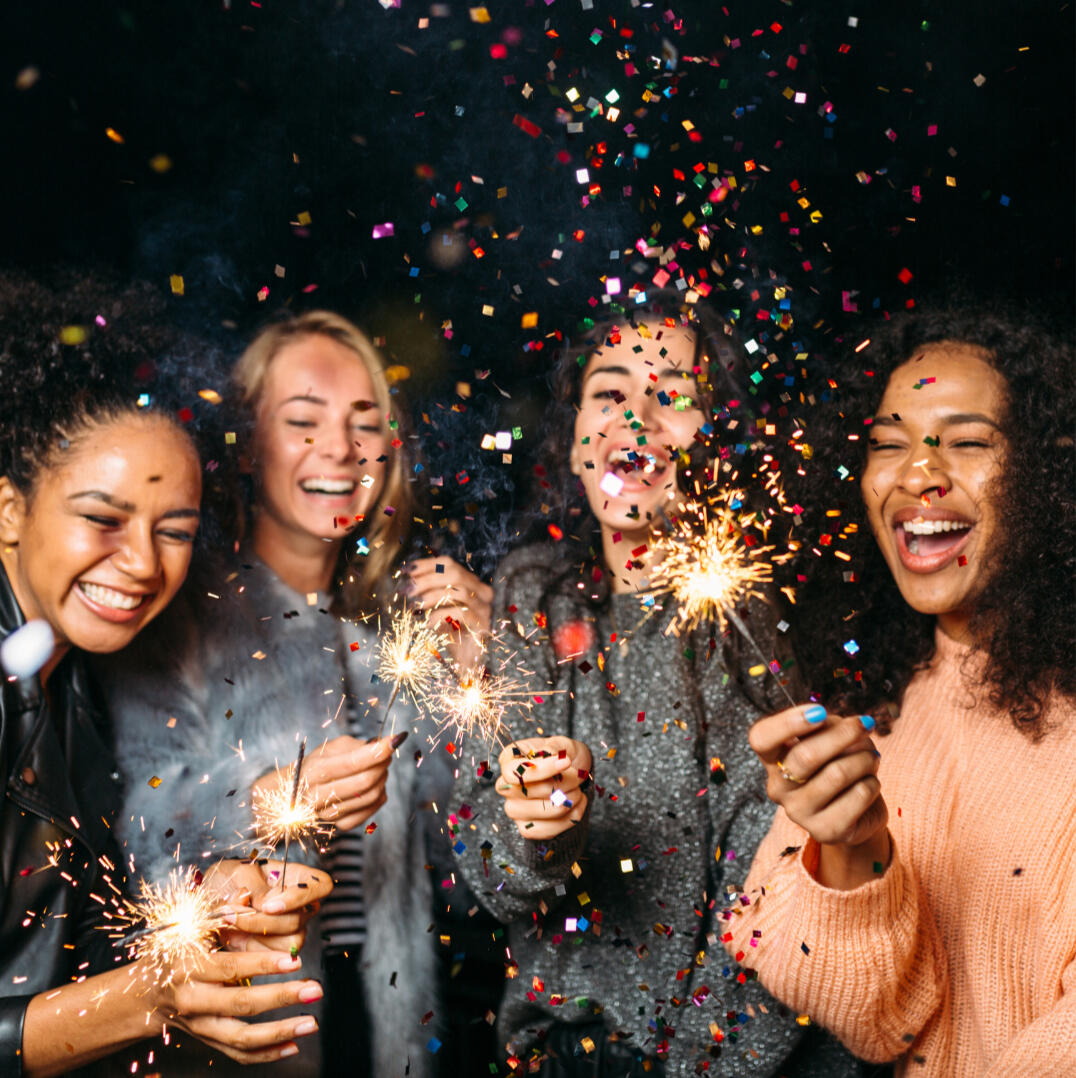 Three female friends smiling and holding sparklers at a nighttime celebration.
